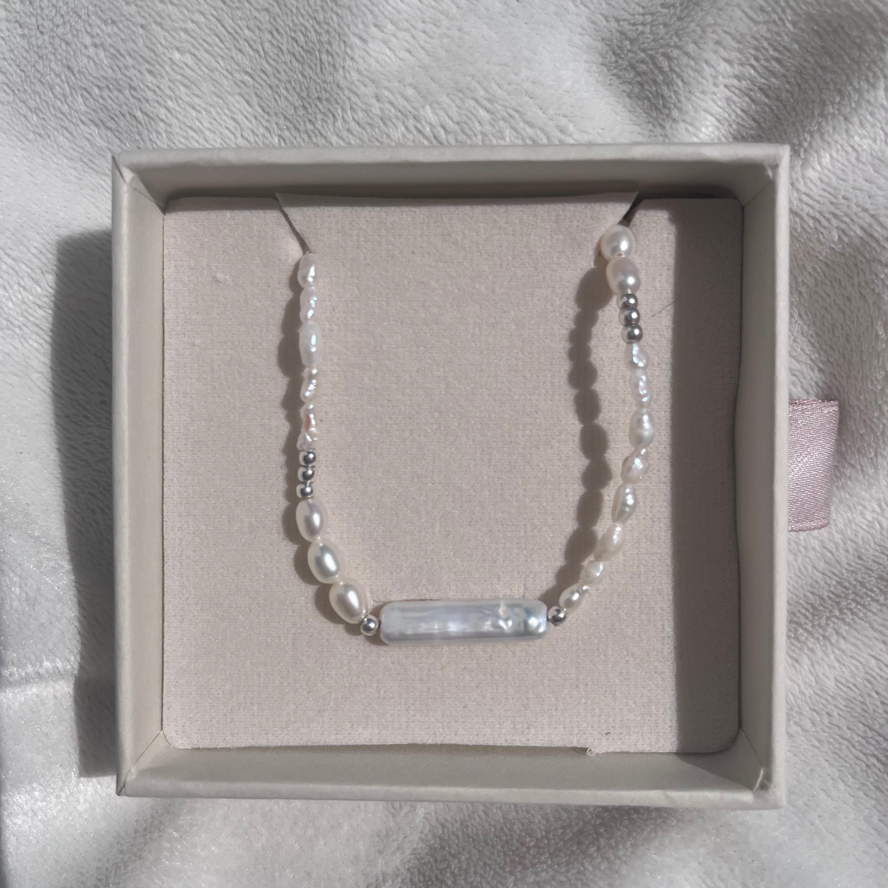 A delicate bracelet with white freshwater pearls and small silver beads, featuring a central white mother-of-pearl bar, displayed inside a light beige jewelry box on a soft white fabric background.