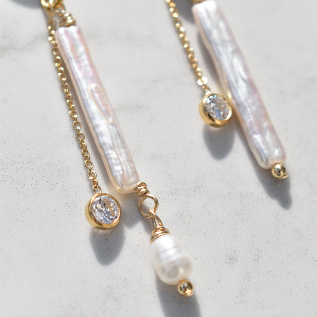 Gold-tone dangle earrings with iridescent white pearl bar beads, small round clear stones, and a dangling pearl bead, shown on a light stone surface.