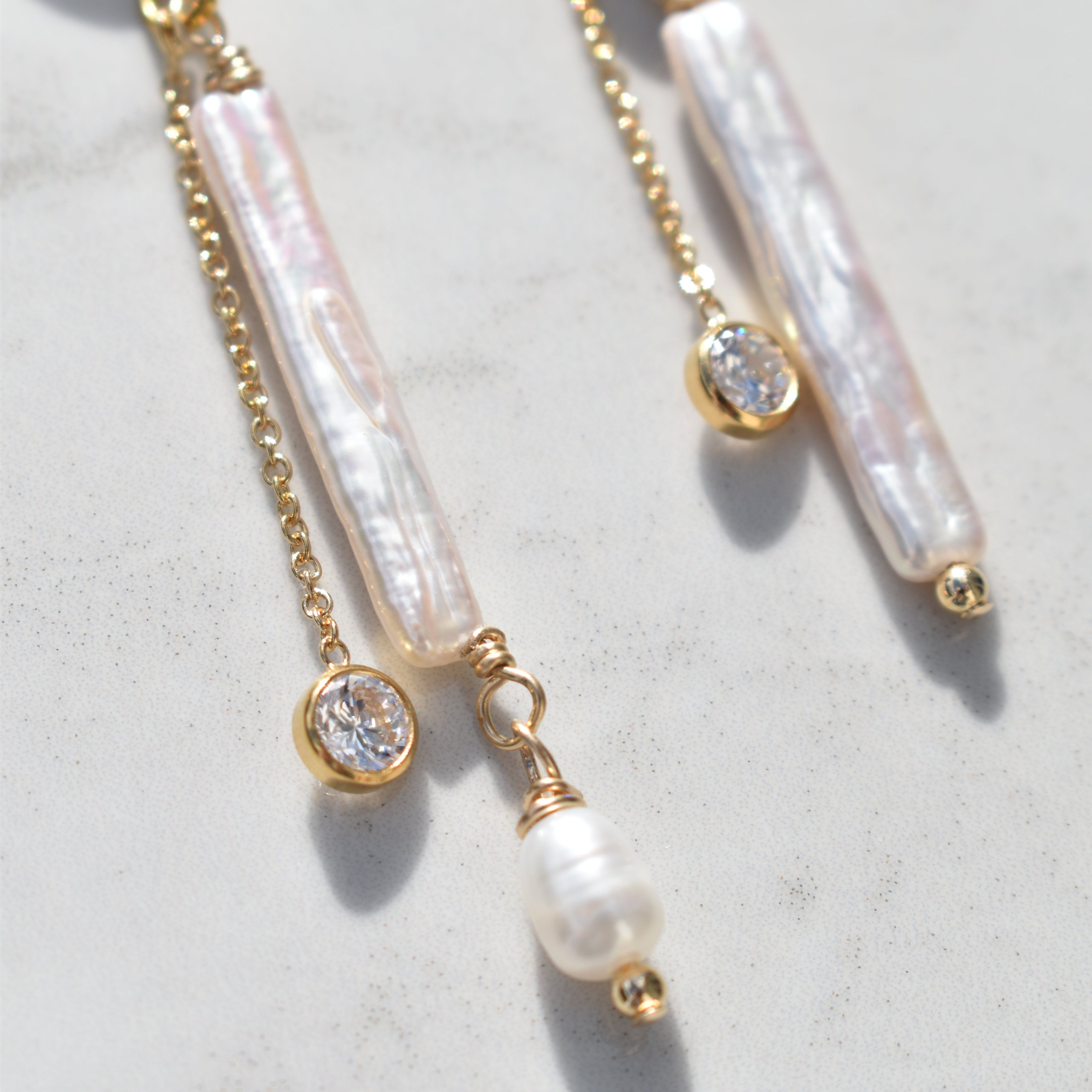 Gold-tone dangle earrings with iridescent white pearl bar beads, small round clear stones, and a dangling pearl bead, shown on a light stone surface.