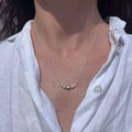 A close-up of a person's neck and upper chest wearing a delicate silver necklace with a curved row of small white pearls, styled with an open white linen shirt.