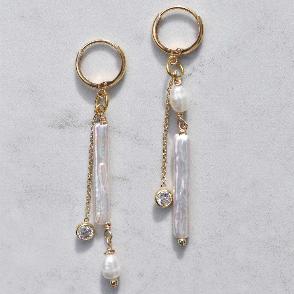 A pair of gold dangle earrings with vertical baroque pearl-like drops, small round pearls, and clear crystal accents, displayed on a light gray marble surface.