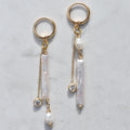 A pair of gold dangle earrings with vertical baroque pearl-like drops, small round pearls, and clear crystal accents, displayed on a light gray marble surface.