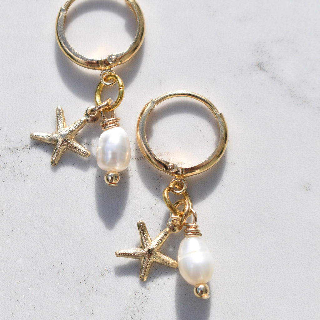 A pair of gold hoop earrings with dangling starfish charms, small gold beads, and white pearl beads, displayed on a light gray marble surface.
