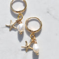 A pair of gold hoop earrings with dangling starfish charms, small gold beads, and white pearl beads, displayed on a light gray marble surface.