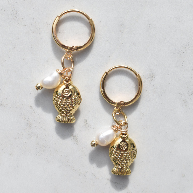 A pair of gold-toned hoop earrings with dangling textured fish charms and small white pearl beads, displayed on a light gray marble surface.