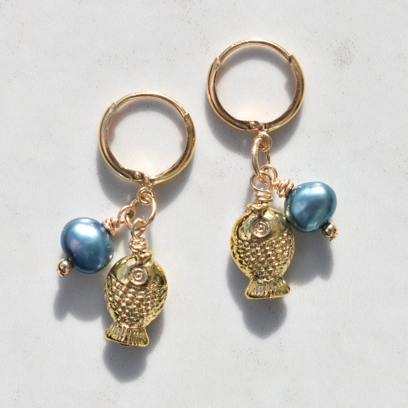 A pair of gold fish-shaped earrings with blue pearl beads, displayed on a light gray marble surface.