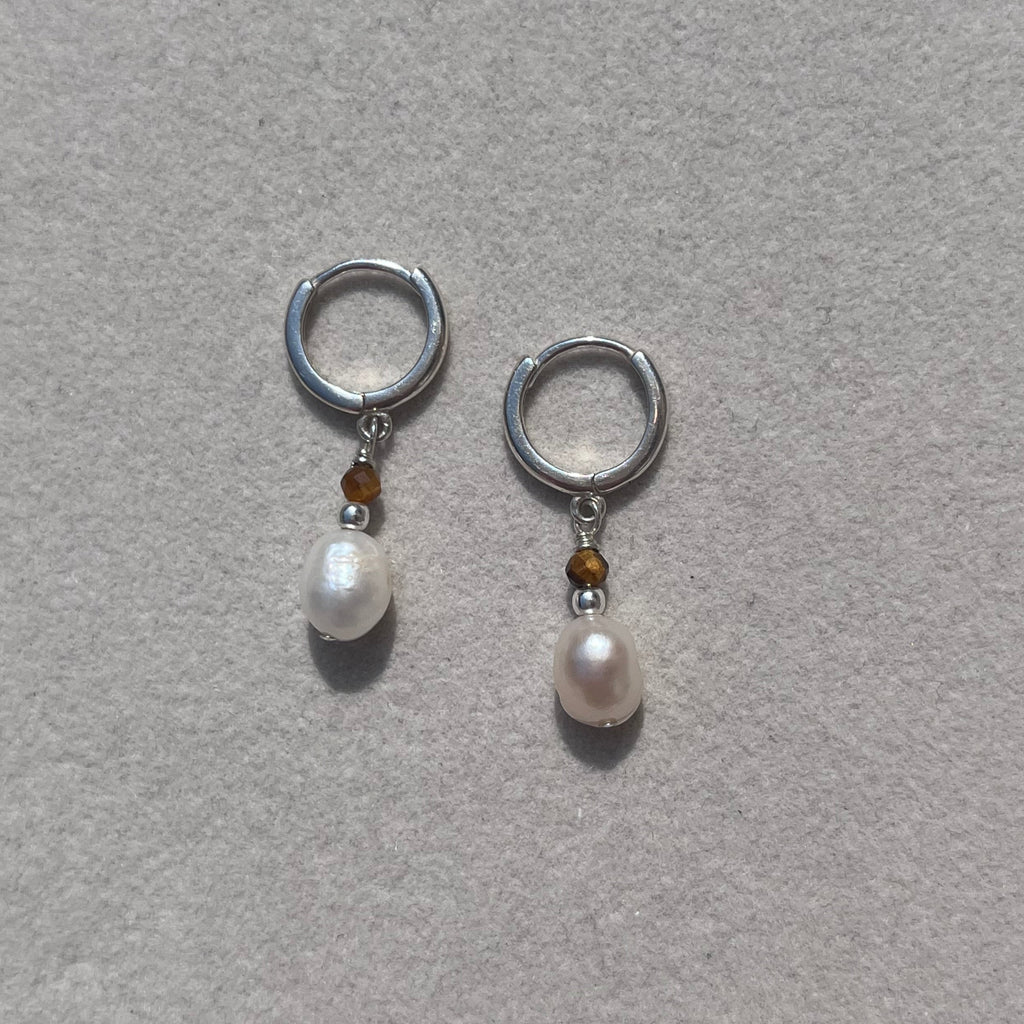 A pair of silver hoop earrings with a small brown bead and a white pearl drop, displayed on a textured light gray surface.