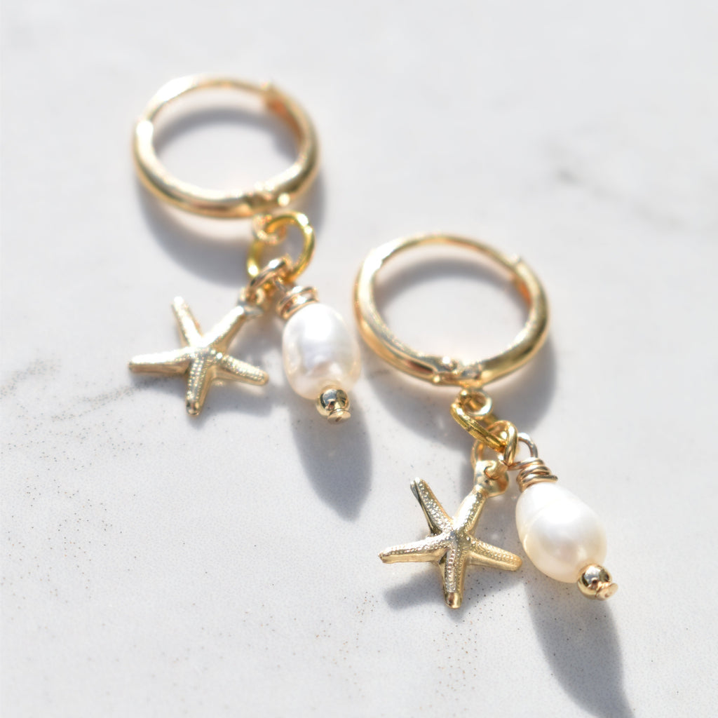 A pair of gold huggie hoop earrings with dangling starfish charms, small clear rhinestone beads, and white pearl drops, displayed on a light marble surface.