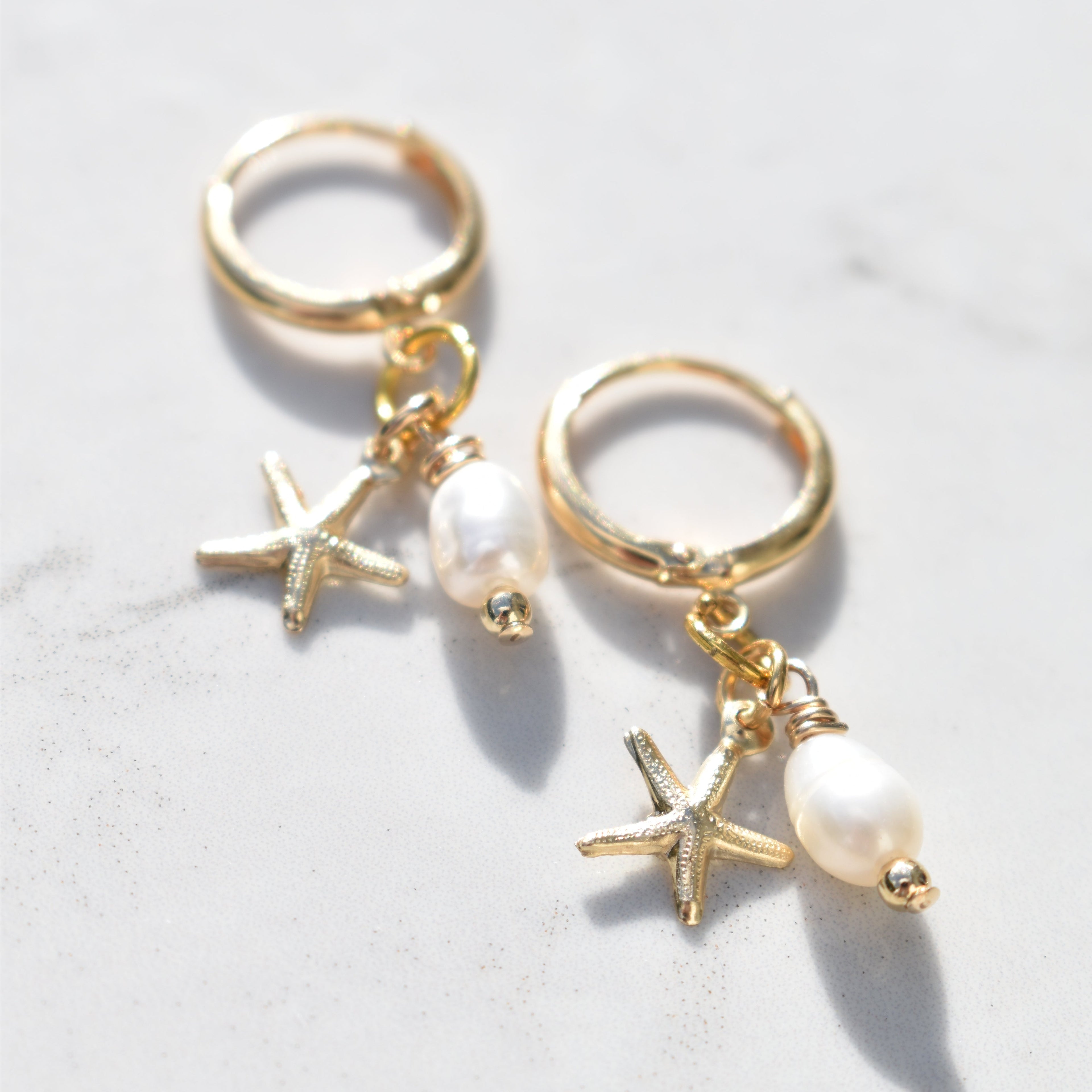 A pair of gold huggie hoop earrings with dangling starfish charms, small clear rhinestone beads, and white pearl drops, displayed on a light marble surface.