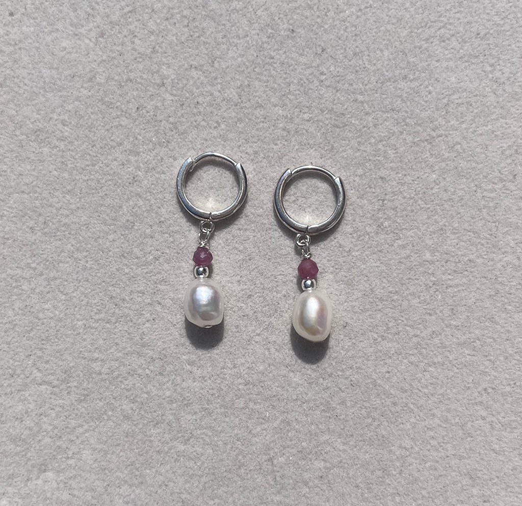 A pair of silver-toned hoop earrings with a small white pearl and a tiny dark red gemstone in the center, displayed on a light gray textured jewelry box.