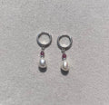 A pair of silver-toned hoop earrings with a small white pearl and a tiny dark red gemstone in the center, displayed on a light gray textured jewelry box.
