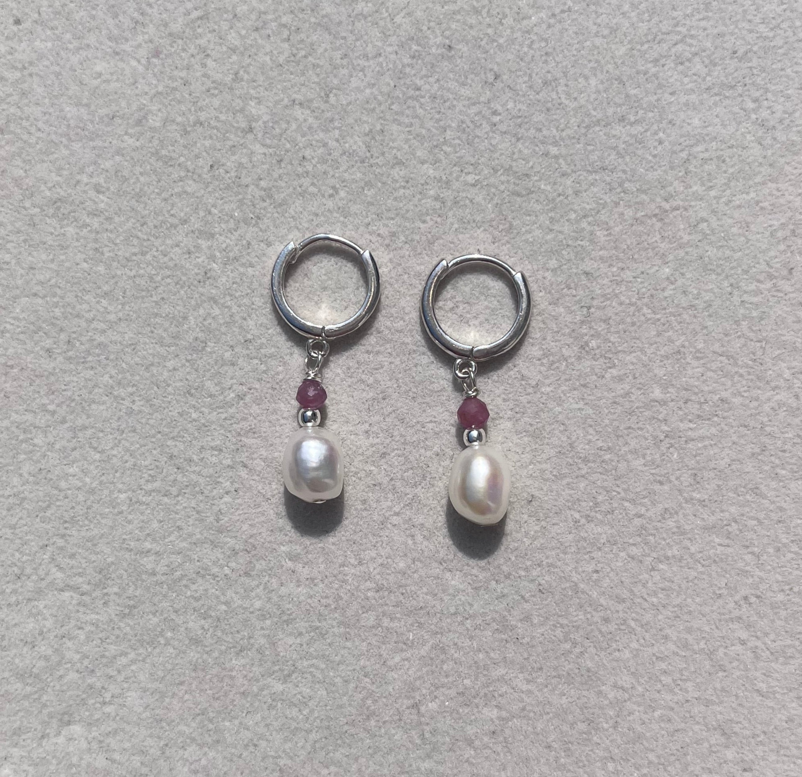 A pair of silver-toned hoop earrings with a small white pearl and a tiny dark red gemstone in the center, displayed on a light gray textured jewelry box.