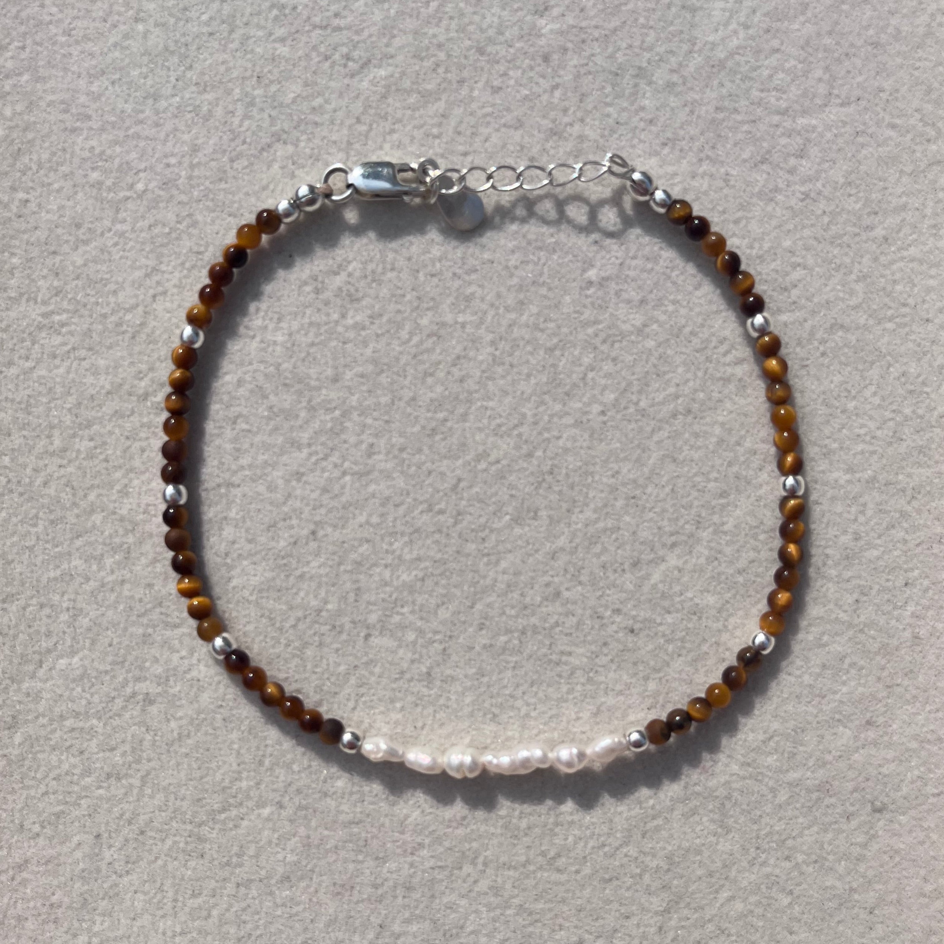A delicate beaded bracelet with brown tiger's eye beads, silver spacer beads, and a silver clasp, displayed on a textured light gray surface.